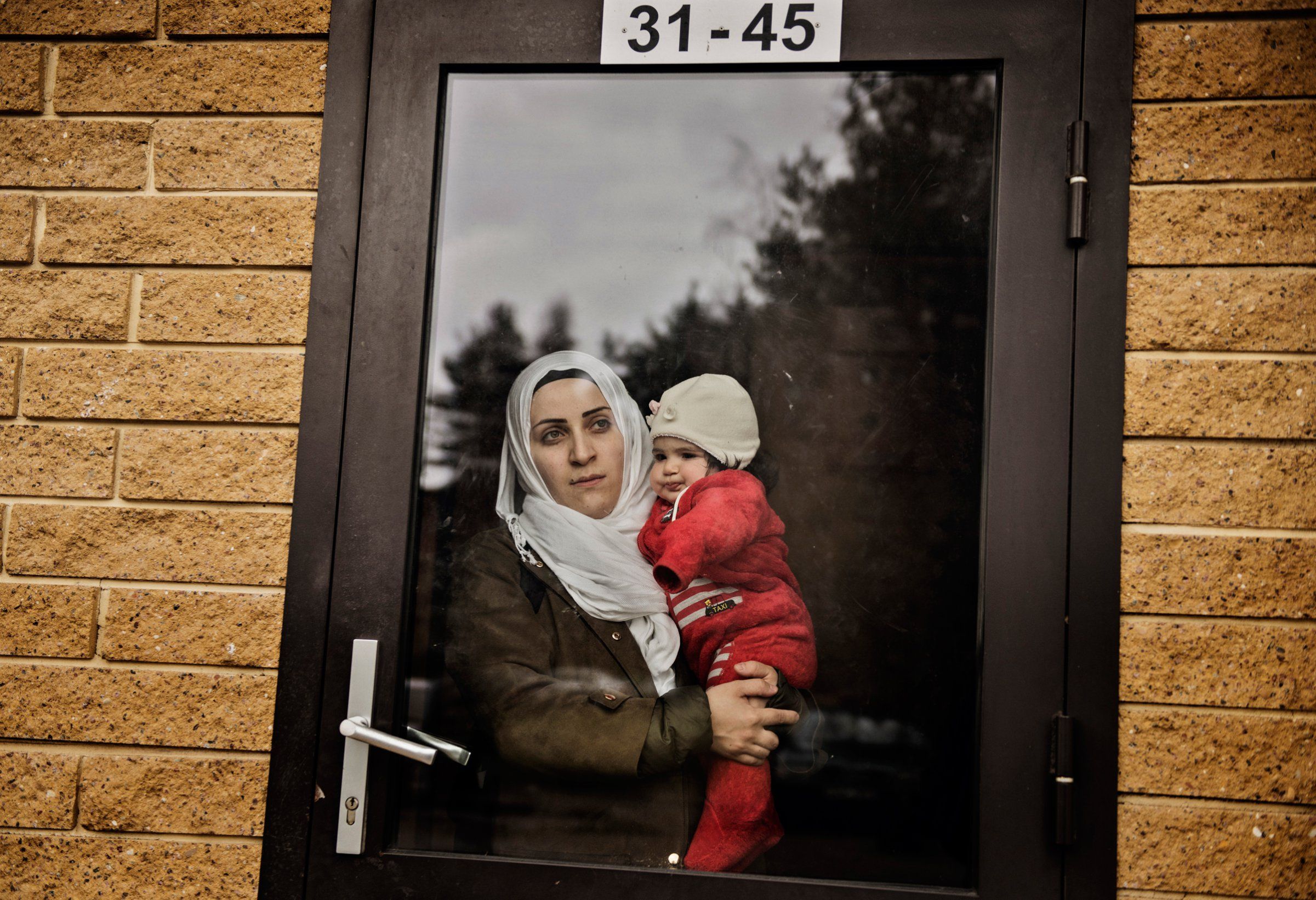 Syrian refugee Taima Abzali stares out the window of her apartment building front door as she holds baby Heln the morning after a long, grueling day of travel and upheaval once again from Athens to their new home in Polva, Estonia. Image by Lynsey Addario. Estonia, 2017. 