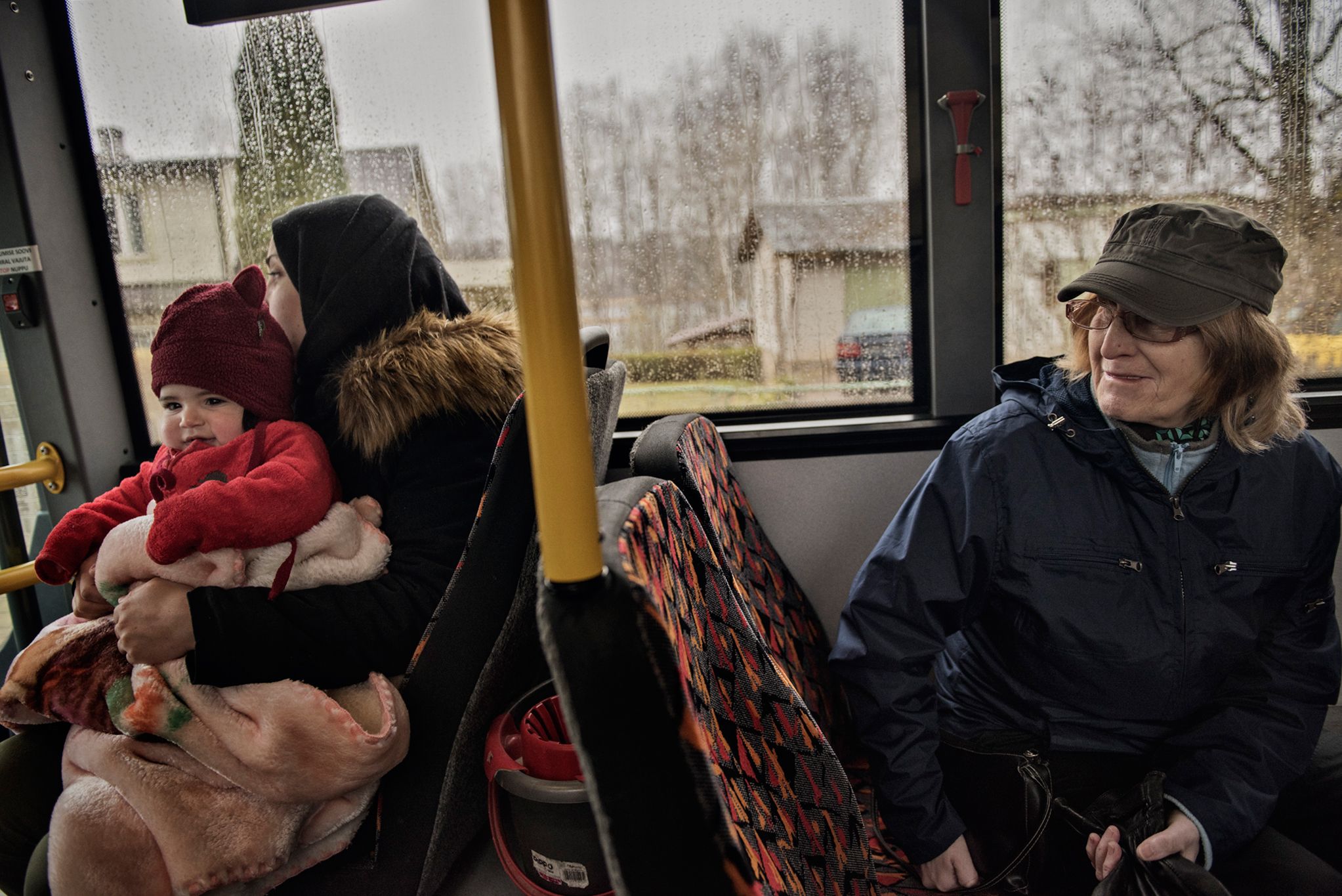 An Estonian woman looks over at Syrian refugees Taimaa Abzali and six month old daughter, Heln, on the bus from Polva to Tartu, where the family will have their orientation with IOM on their second full day in Estonia. Image by Lynsey Addario. Estonia, 2017. 
