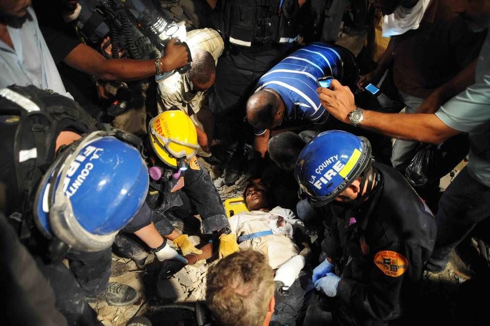 Roger Sainfort (in striped shirt) checks on his wife, Ginette Sainfort, while members of the Los Angeles County Fire Department Search and Rescue Team give her an IV following her rescue from the parking lot of a collapsed three-story bank. She spent nearly a week buried in rubble. Image by Petty Officer 2nd Class Justin S / U.S. Navy. Haiti, 2010.