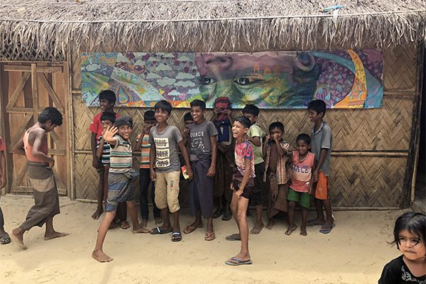 In August, children at a Child-Friendly Space in Balukhali camp, in Bangladesh, pose in front of a section of an Artolution mural sent from New York as a message of support to Rohingya refugees. Image by Jaime Joyce. Bangladesh, 2018.