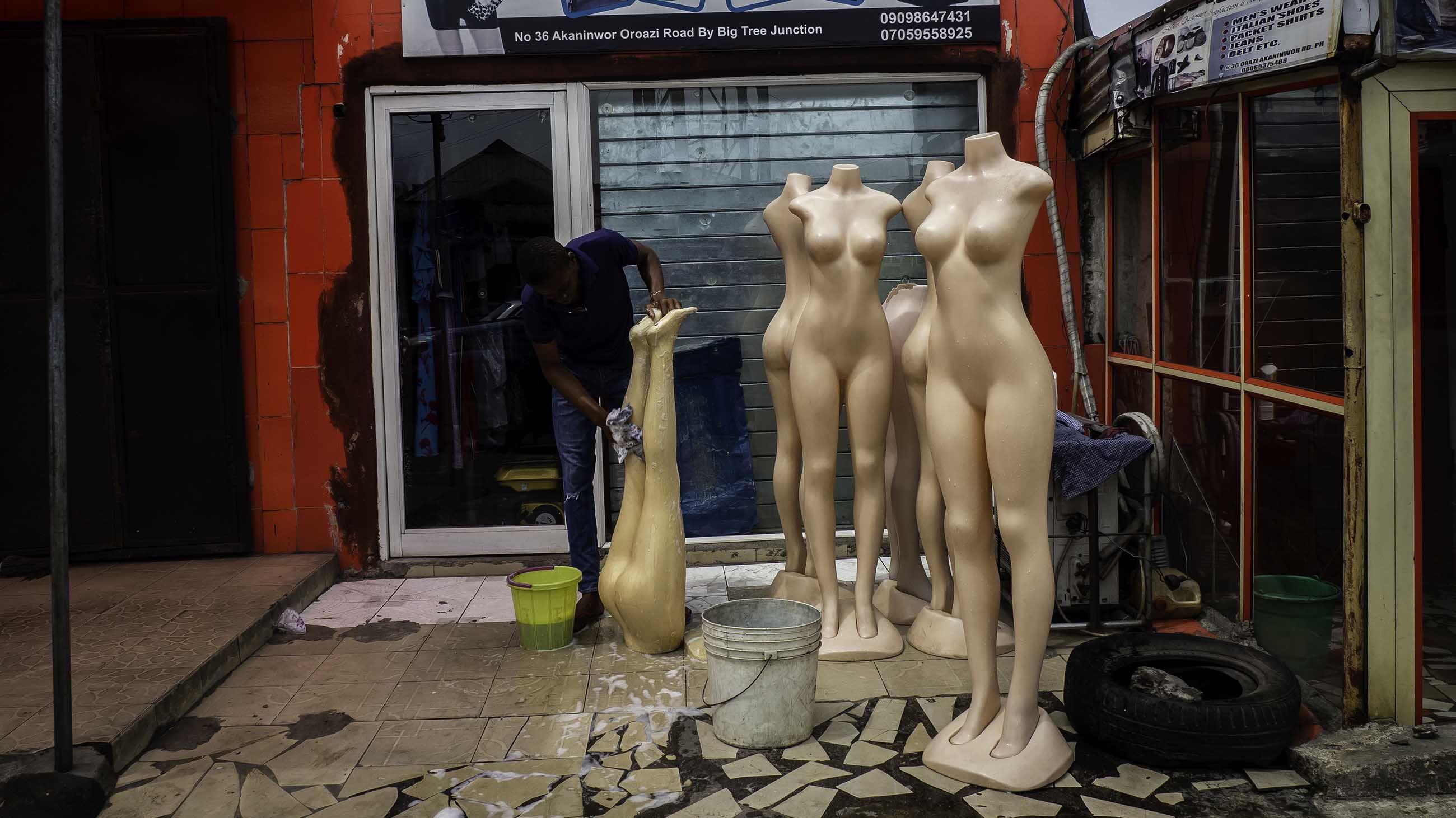 Soot covers everything—buildings, plants, and people. Here, a shop keeper wipes soot off mannequins in front of his fashion store. Image by Larry C. Price. Nigeria, 2018.