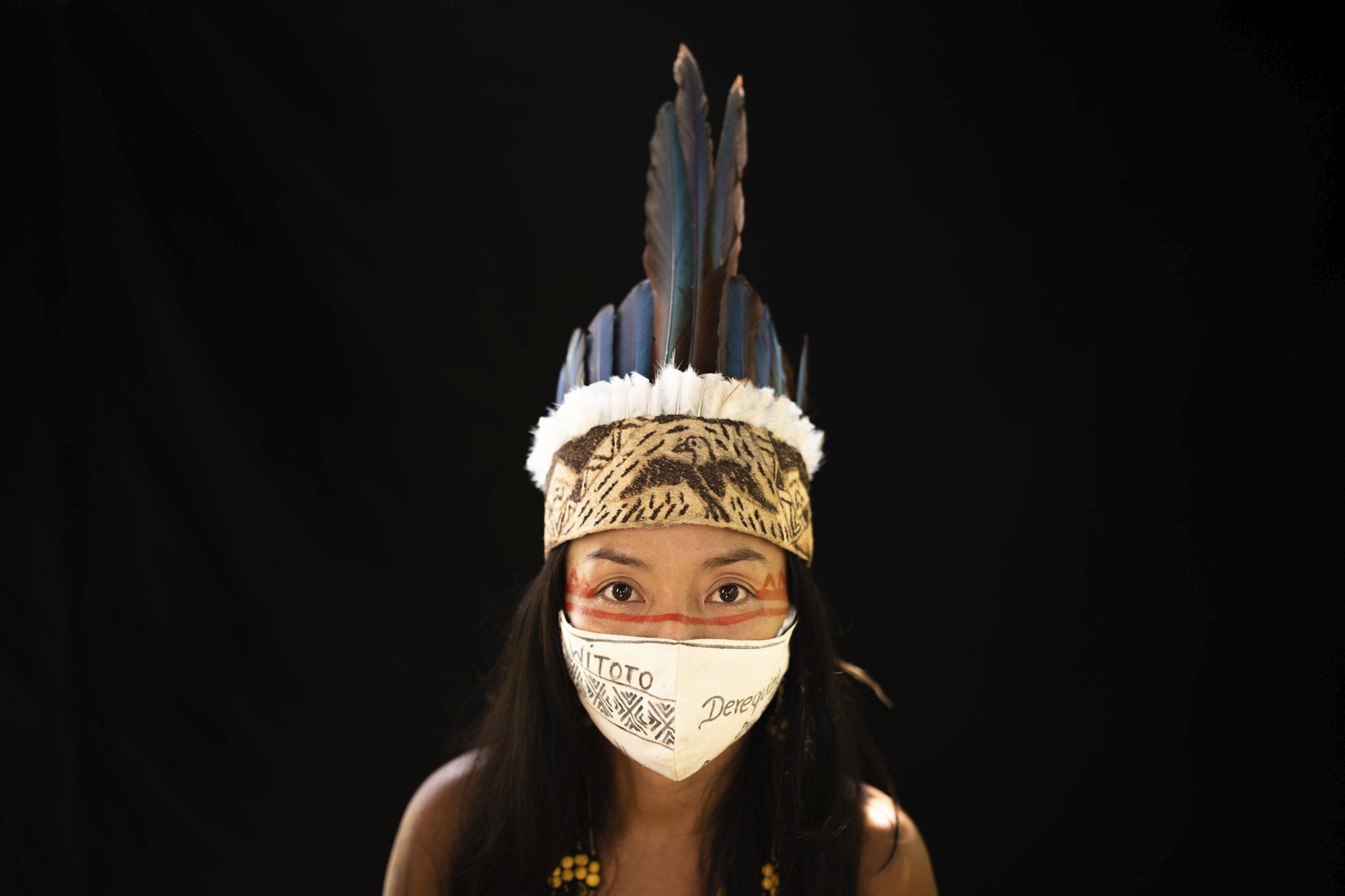 Vanda Ortega, 33, of the Witoto indigenous ethnic group, poses for a portrait wearing the traditional dress of her tribe and a face mask amid the spread of the new coronavirus in the Park of the Tribes community, Manaus, Brazil, Sunday, May 31, 2020. Ortega has become one of the symbols of the indigenous fight against the pandemic after raising her voice to denounce government neglect, including the undercounting of indigenous patients who were, instead, registered as multiracial in the public health system. Image by Felipe Dana / AP Photo. Brazil, 2020.