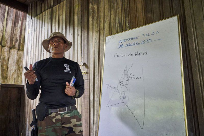 José Gregorio, wearing camo pants, a long sleeve shirt, and a bucket hat, holds several markers as he shows his team the plan written on a whiteboard with the route to be carried out in the routine inspection of his territory before leaving.