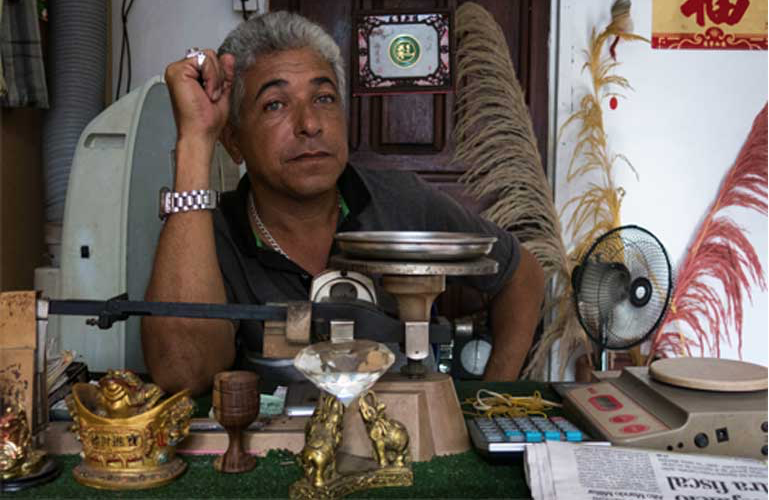A Tumeremo gold pawn shop owner sits behind his desk while enjoying the luxury of a cooling desk fan. He says that he is happy to buy and sell gold, and that it is far too dangerous to work in or around the mines. Image by Bram Ebus. Venezuela, 2017.
