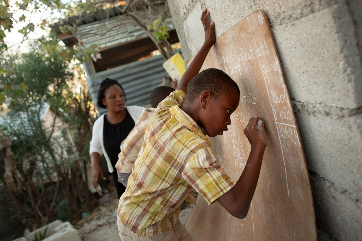 Baron Ritcharmant, 9, right, and his brother Celestin Shneider, 7, with the encouragement of their mother Edrice Elvige, write out their math homework in chalk in the backyard of the family's home in the Canaan 4 neighborhood. An active community organizer since she moved to Canaan in 2010, Elvige dreams of a center where the neighborhood children can go for activities and basic health care. Image by Allison Shelley. Haiti, 2019.