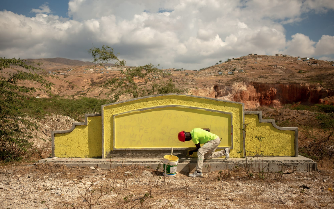 A worker paints over graffiti on an entrance sign to the St. Christophe memorial. An elaborate monument has been built further up the road but rarely receives visitors outside of the occasional international group. Image by Allison Shelley. Haiti, 2019.