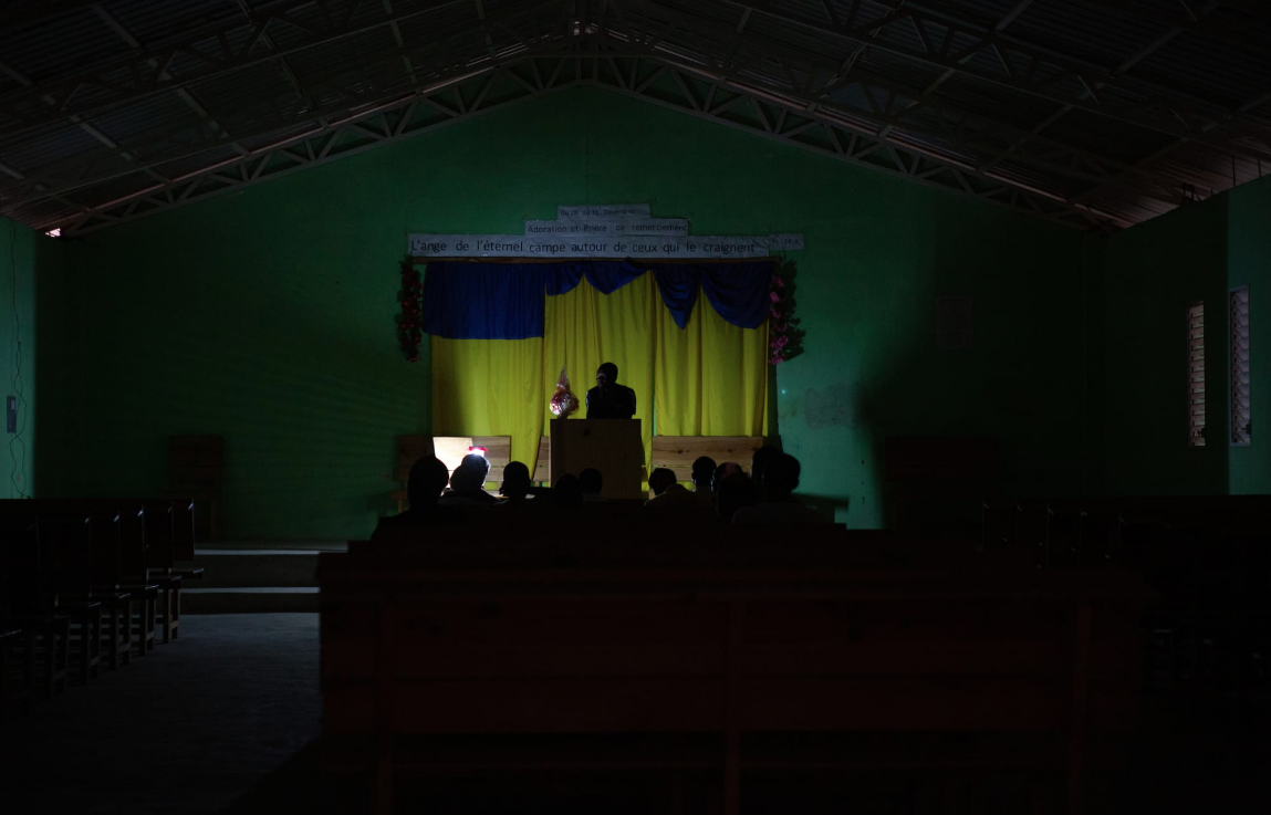 A preacher gives a sermon by the light of a solar lamp at Eglise Baptiste TBM de Titanyen, a Baptist church. This section of Canaan is without electricity, pirated or legitimate. Image by Allison Shelley. Haiti, 2019.