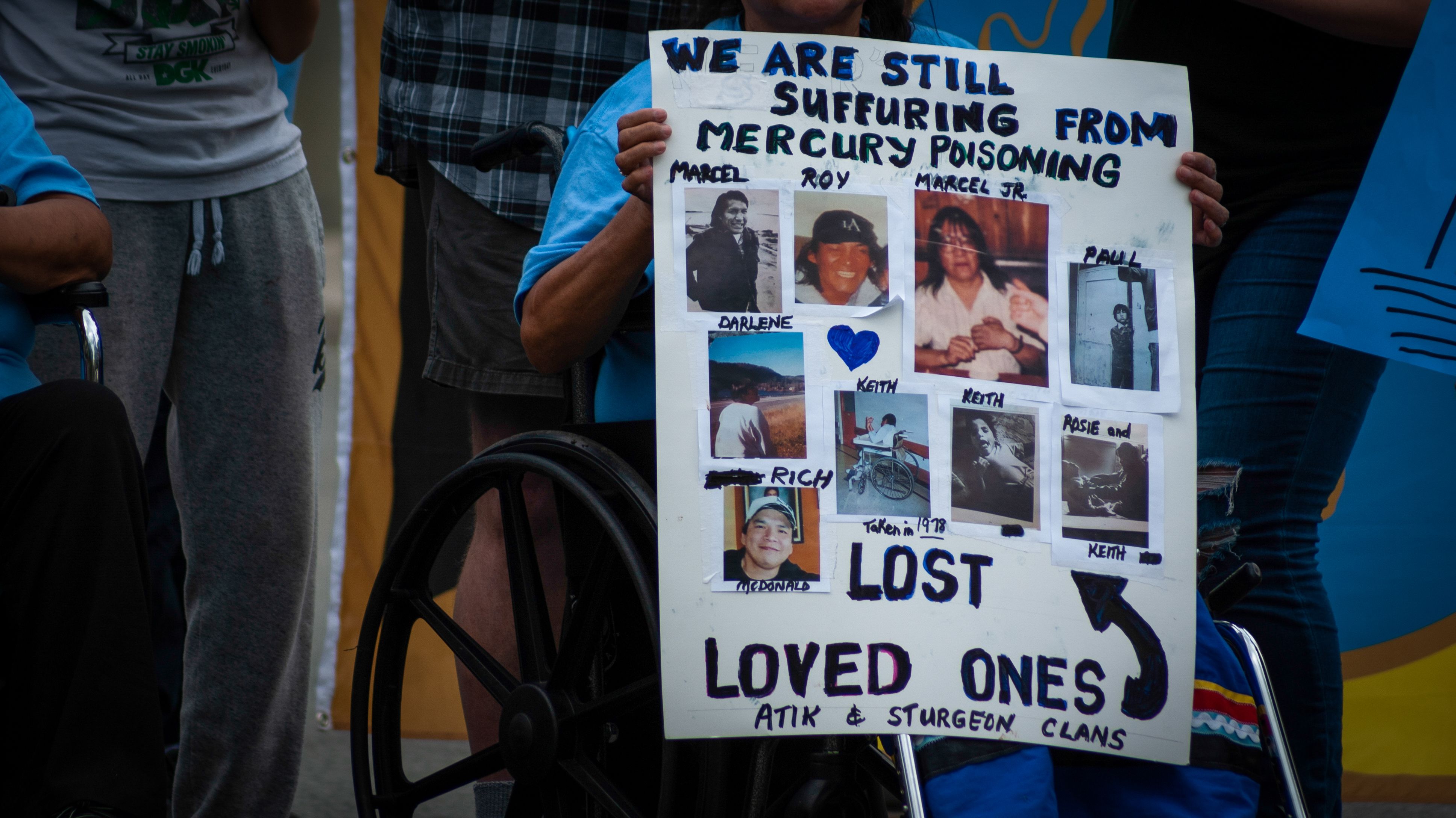 A woman sits in a wheelchair and holds a sign which reads, "We are still suffering from mercury poisoning." It also features photos of family members who have passed away.