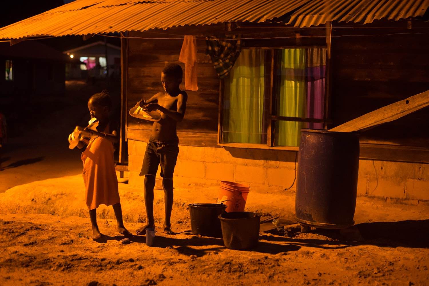 Children clean their shoes for school in the orange glow of a street light on Wednesday, March 15, 2017 in the village of Adjuma Kondre. It will take Alcoa years to repair thousands of acres of mined land around the village. Image by Stephanie Strasburg. Suriname, 2017.
