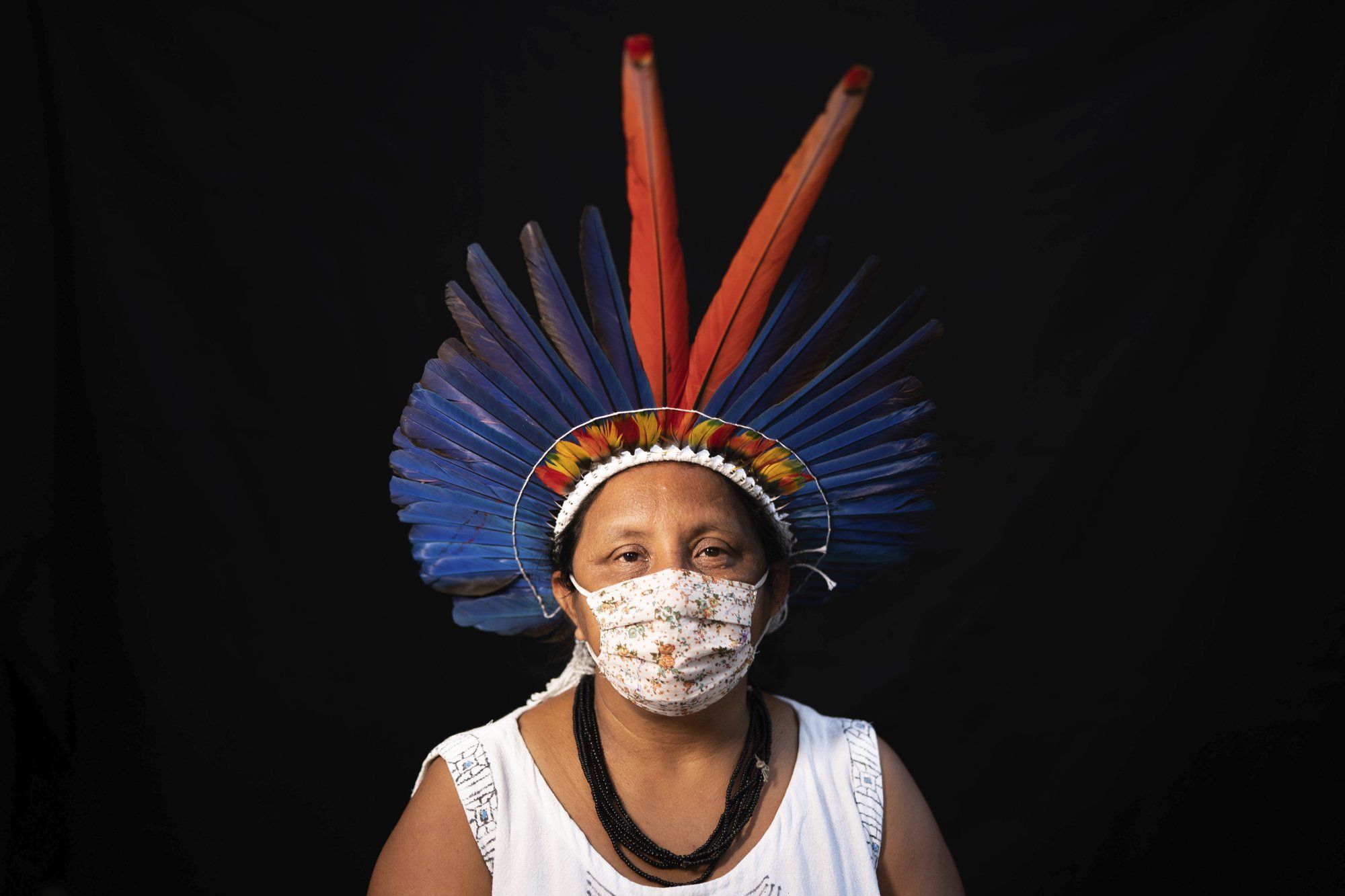 Sonia Vilacio, 46, of the Sateré Mawé indigenous ethnic group, poses for a portrait wearing the traditional dress of her tribe and a face mask amid the spread of the new coronavirus in Manaus, Brazil, Wednesday, May 27, 2020. The new coronavirus has hit Sônia Vilacio twice: when tourists disappeared from Manaus and she was left without an income from selling arts and crafts, and soon after when she fell ill with a fever, shortness of breath, and a cough, telltale signs of COVID-19. Image by Felipe Dana / AP Photo. Brazil, 2020.