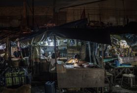 A woman works on bookkeeping using the light of her phone in the Market 3 slum in the Navotas Fish Port Complex. Market 3 has electricity but no plumbing. Residents often turn to drugs to alleviate hunger and stay awake for the long hours they work at low pay. Image by James Whitlow Delano. Philippines, 2017.